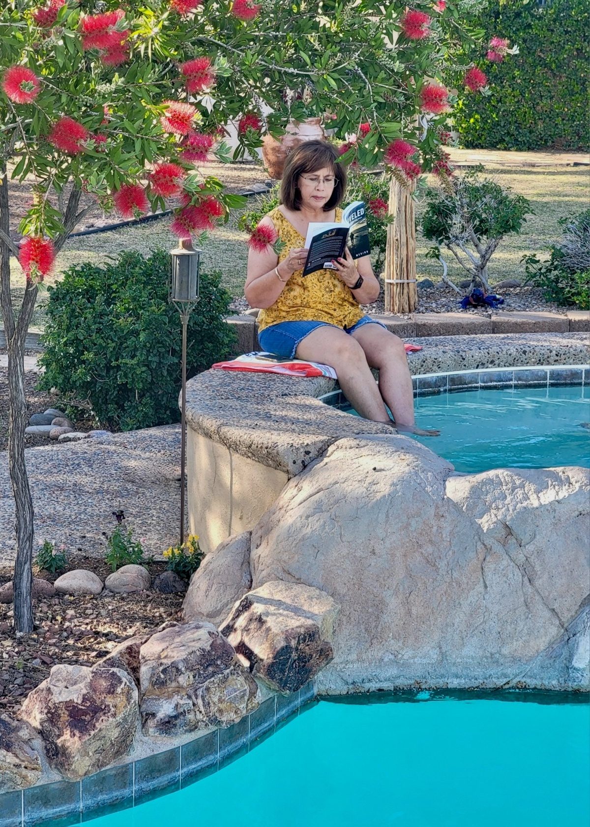 Sylvia reading Unraveled on the edge of a pool under a flowering tree